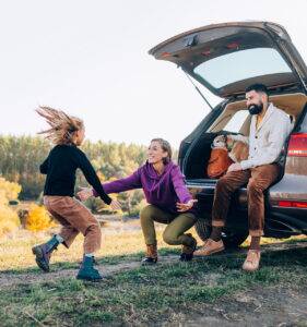 Man sitting in car with dog while woman and child play nearby