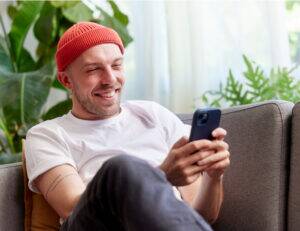 Young man in red beanie smiling while texting on smartphone from gray couch